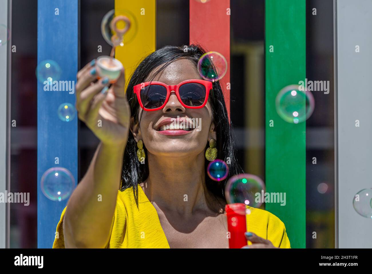 Vorderansicht einer erwachsenen Latina Frau lächelnd und hält Blasen Zauberstab in einem gelben Kleid und Sonnenbrille vor einem Hintergrund von bunten Bars auf einem sonnigen Stockfoto