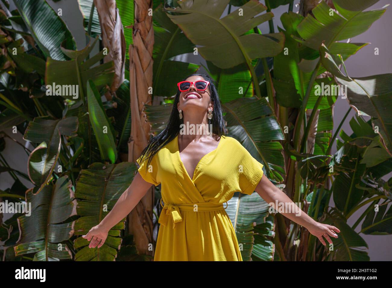 Vorderansicht einer Latina-Frau, die in einem gelben Kleid und einer Sonnenbrille steht und mit ausgestreckten Armen auf einem Hintergrund mit grünen Pflanzen auf einem sonnigen Blick nach oben blickt Stockfoto