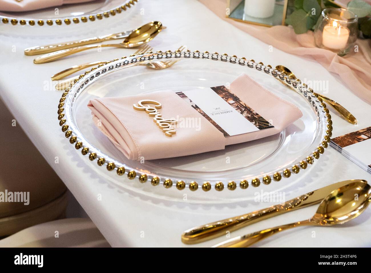 Goldfarbenes Besteckbesteck mit voller Tischeinstellung neben goldgerandigem Glasteller als Teil einer Tischeinstellung im Hochzeitsstil mit pinkfarbener gefalteter Serviette und einem Namen des Gastes auf der Oberseite. Stockfoto