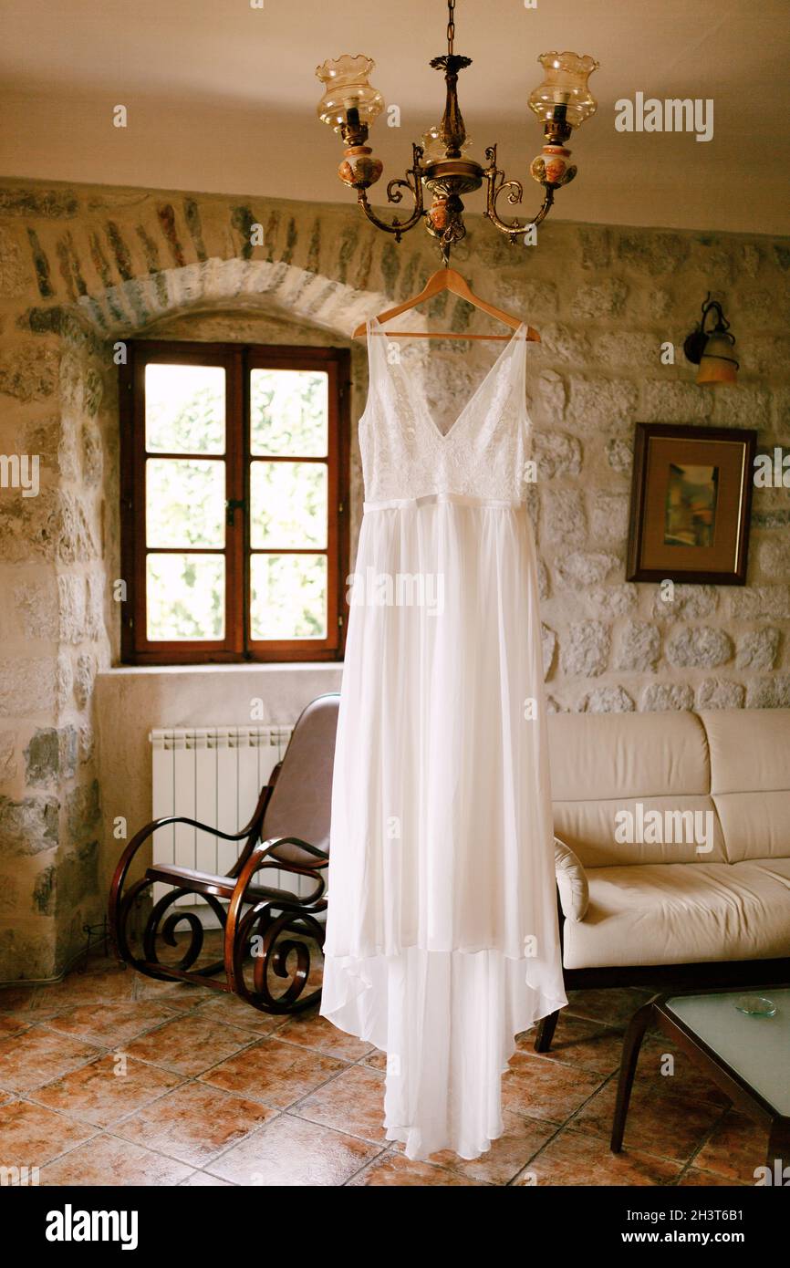 Budva, Montenegro - 21. august 2020: Hochzeitskleid der Braut auf einem Holzbügel auf einem Kronleuchter in einem Raum mit Sofa und einem Zimmer Stockfoto
