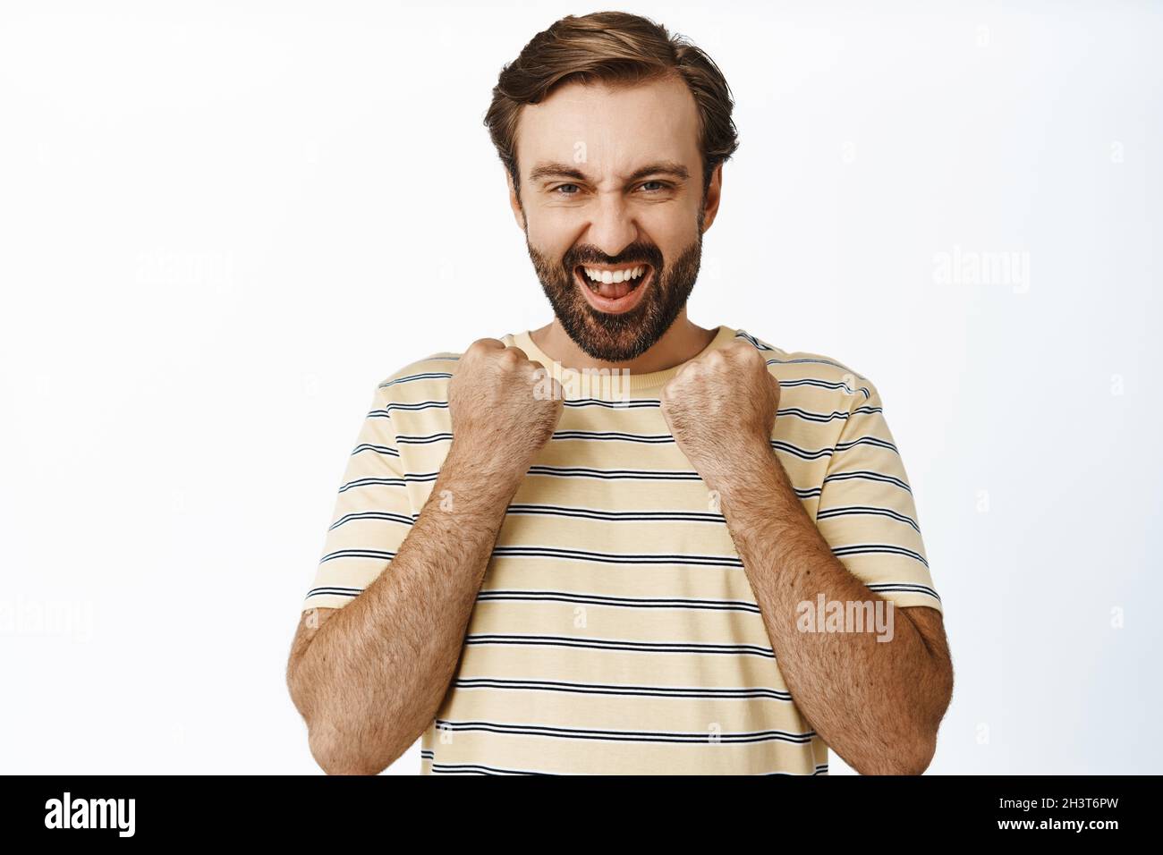 Enthusiastischer bärtiger Mann klatscht mit den Fäusten, lacht und lächelt zufrieden, sieht ermutigt und selbstbewusst aus und steht auf weißem Hintergrund Stockfoto