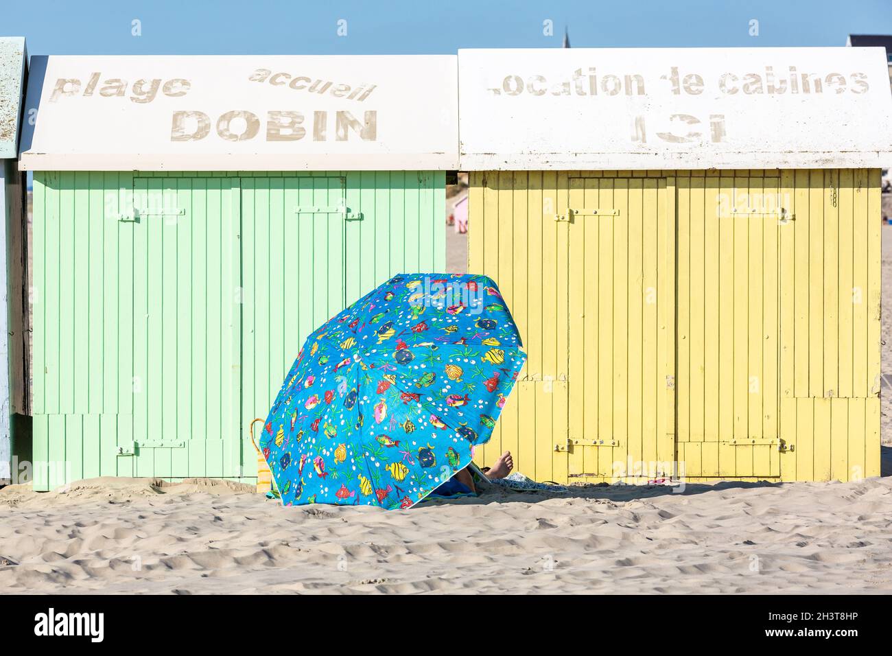 Person, die unter einem blauen Sonnenschirm in der Nähe von bunten Strandhütten ruht. Berck-Plage, Opalküste, Frankreich Stockfoto