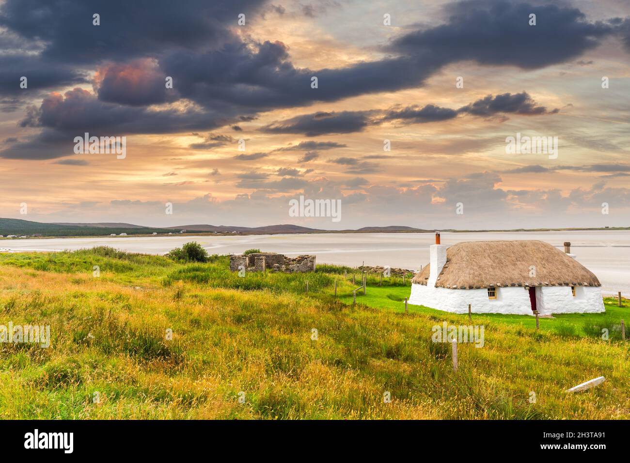 Traditionell gebaute weiße Hütte mit Strohdach, neben der türkisfarbenen Bucht, mit stürmischen bewölkten dunklen Himmel über.Insel North Uist, Schottland Stockfoto
