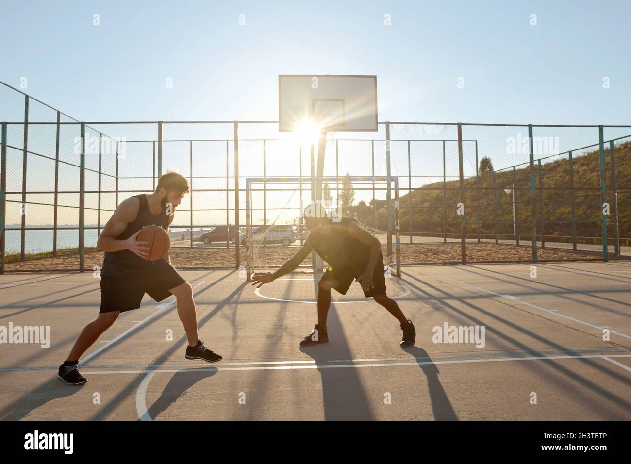 Junge Sportler spielen Basketball auf dem Sportplatz. Schwarze und europäische männliche Freunde tragen Sportswear und Sneakers. Basketballspieler in der Stadt. Meer beh Stockfoto