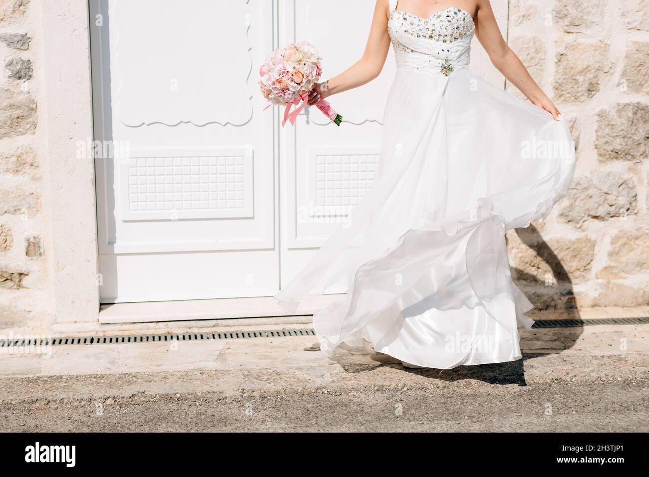 Eine Braut in einem stilvollen Brautkleid und mit einem brautstrauß steht an der schönen weißen Tür von an Altes Steinhaus Stockfoto