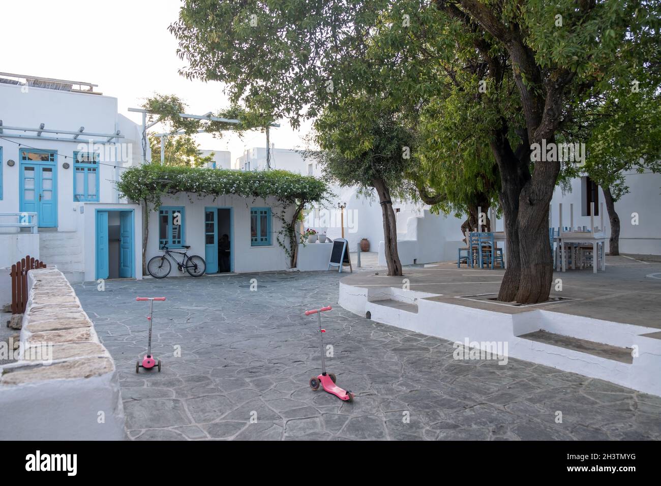 Griechenland, Insel Folegandros. Zwei Kinderroller in der Kopfsteinpflasterstraße, traditionelle Gebäude und Bäume am Chora-Stadtplatz. Kykladen. Stockfoto