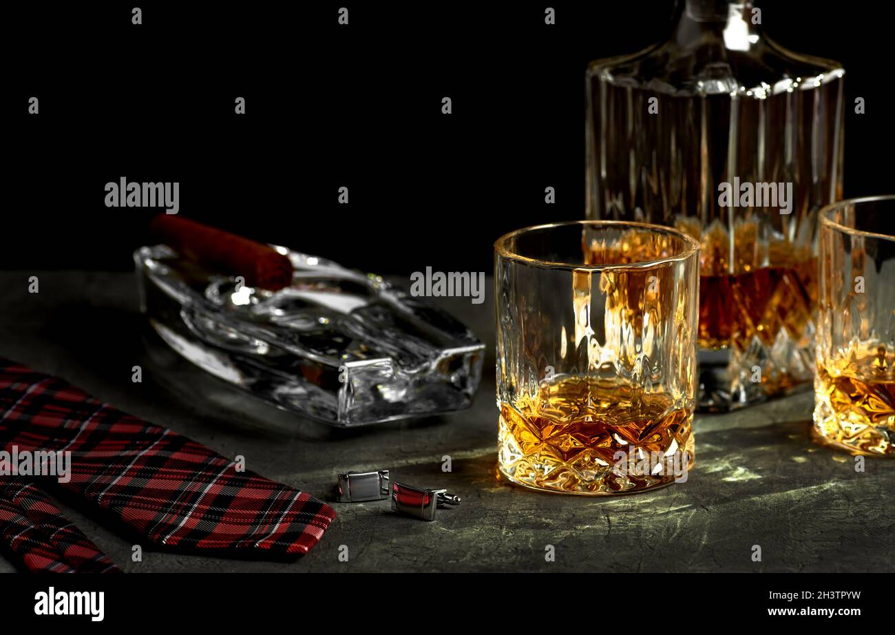 Stillleben mit einer Flasche und zwei Gläsern Whisky auf einer Steinplatte in einer Bar. Kubanische Zigarre in einem Aschenbecher und eine rote Krawatte auf schwarzem Hintergrund - Spa Stockfoto