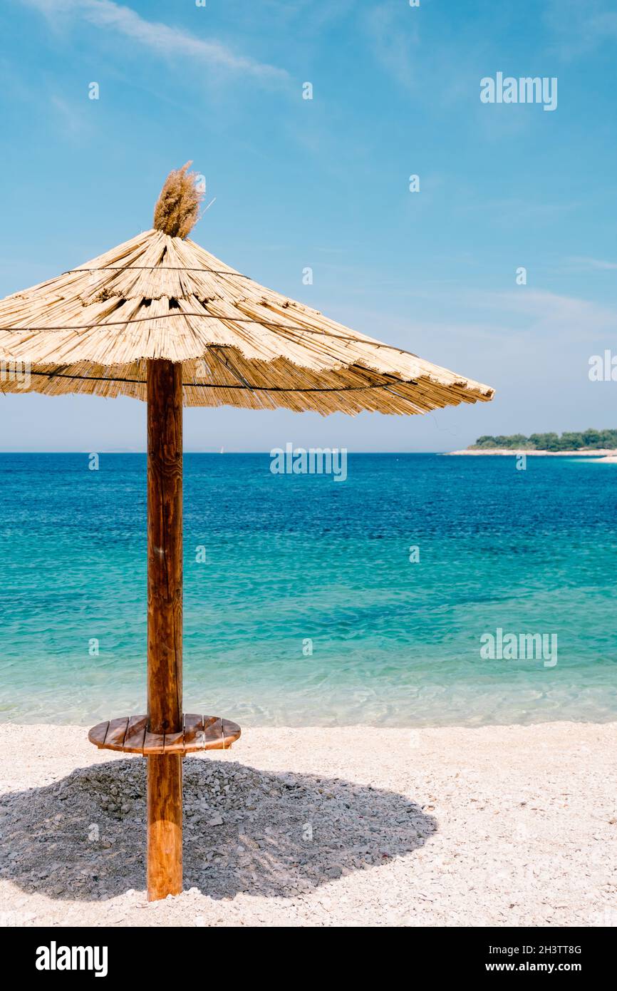 Ein Strand mit Strohschirm gegen den blauen Himmel und azurblauem Wasser an einem Sandstrand in Kroatien, in der Stadt Primosten. Stockfoto