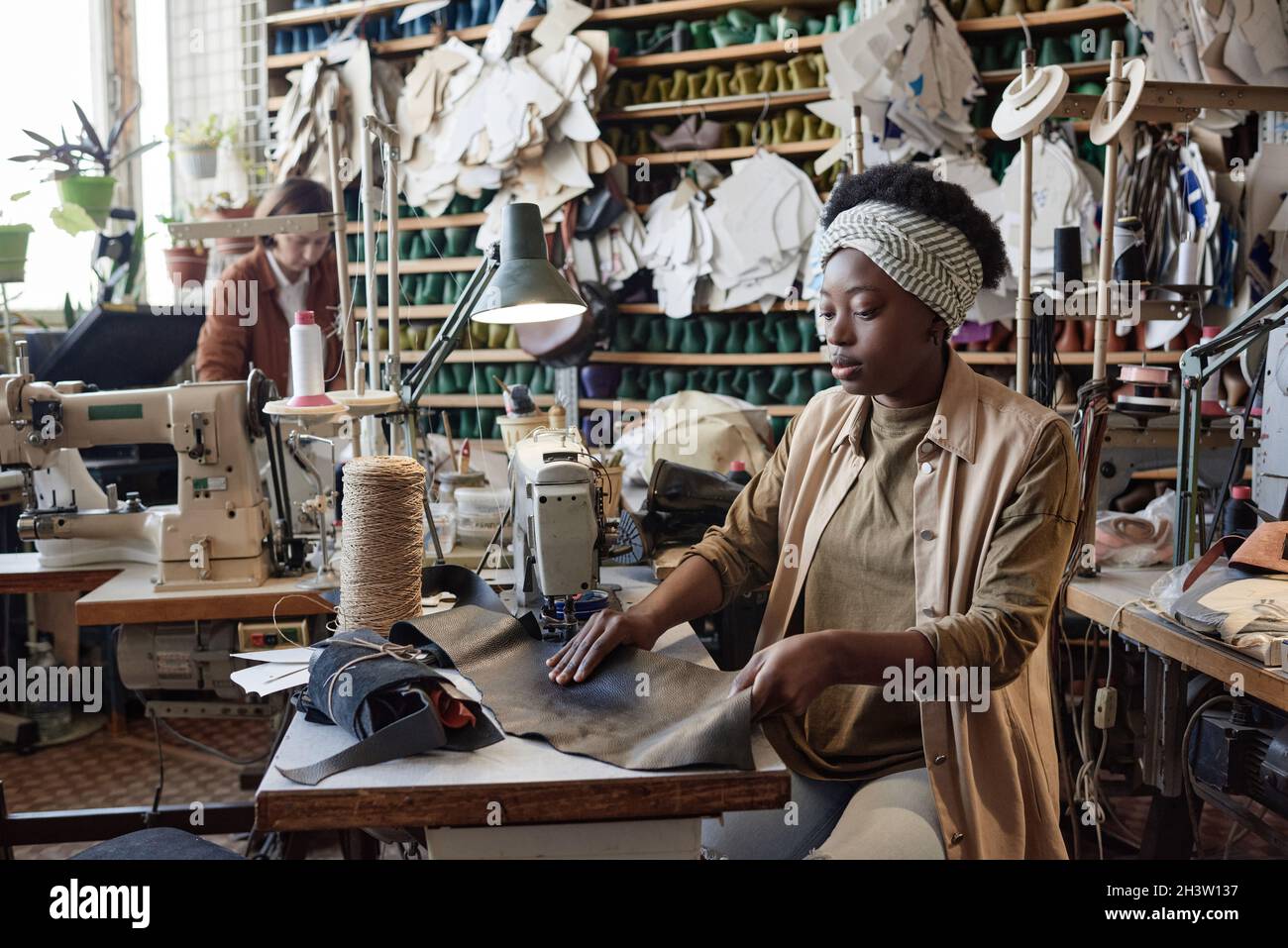 Afrikanische Schneiderin sitzt an ihrem Arbeitsplatz mit Nähmaschine und Nähprodukten in der Fabrik Stockfoto