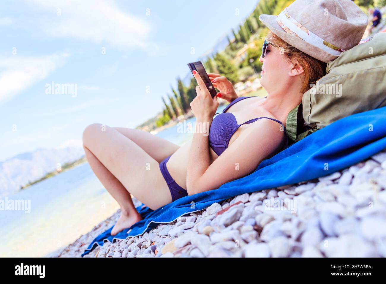 Smartphone am Strand: Schönes junges Mädchen mit Strohhut im Bikini liegt am Kiesstrand, Urlaub in italien Stockfoto