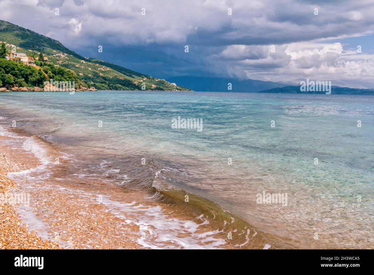 Wunderschöne Landschaft - türkisfarbenes Meerwasser, goldener Sand, grauer Himmel mit dunklen, stürmischen Wolken und Bergen am Horizont. Stockfoto