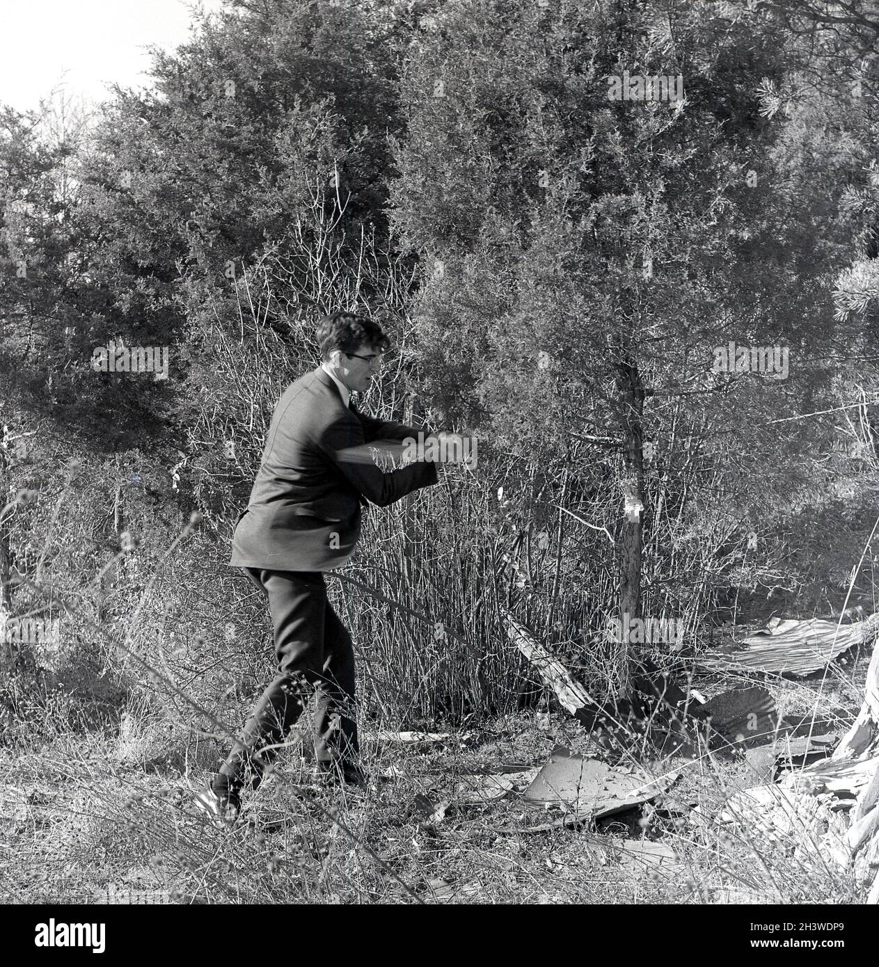 1968, historisch, sommerlich und draußen in einem Wald oder Waldgebiet, ein junger Mann in einem Anzug hackt einen kleinen Baum mit einer Axt, Middletown, Virginia, USA. Stockfoto