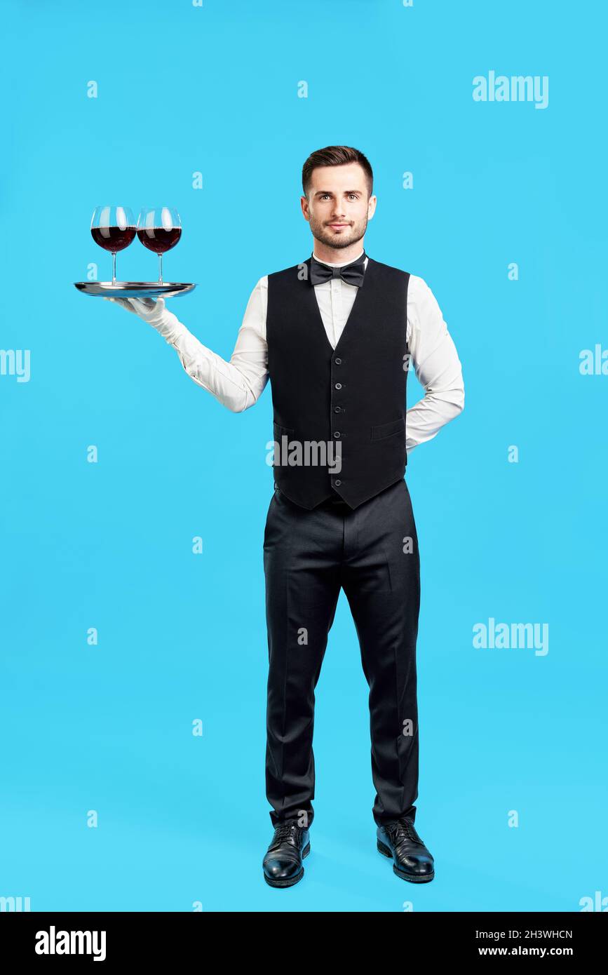 In voller Länge Porträt eines eleganten männlichen Kellners mit Gläsern Wein auf blauem Hintergrund Stockfoto