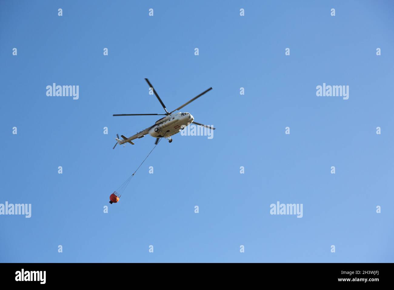 Hubschrauber fährt in den Feuerbereich, um Waldbrände zu löschen. Helikopter mit Wassereimer am blauen Himmel. Stockfoto