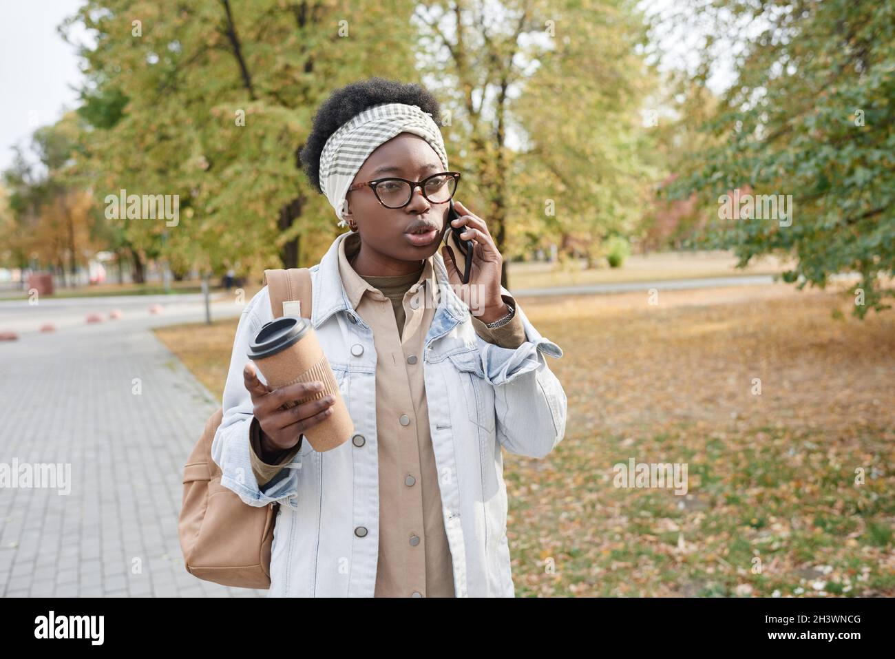 Afrikanische junge Frau, die während ihrer Kaffeepause im Park spazierengeht und mit dem Handy spricht Stockfoto