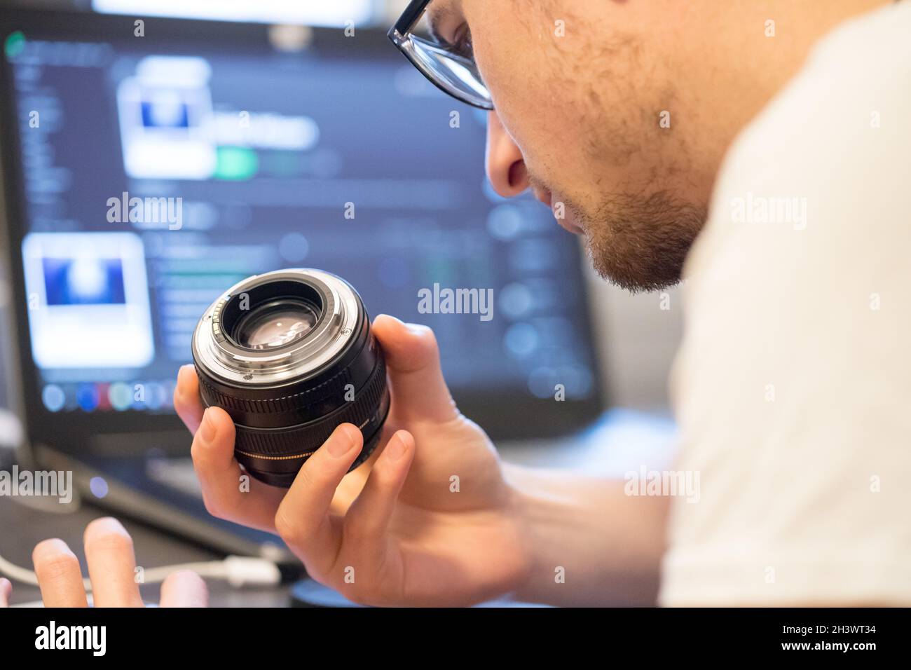 Der männliche Fotograf hat ein Objektiv in der Hand und schaut darauf. Laptop im unscharfen Hintergrund. Stockfoto