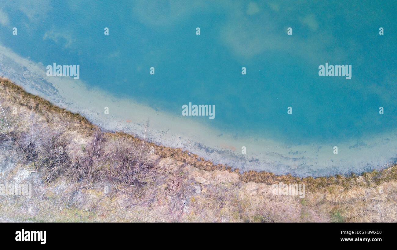 Luftaufnahme eines malerischen Ortes, an dem im Frühling das durchsichtige türkisfarbene Wasser eines Waldsees auf ein steineres Ufer mit Bäumen trifft. Stockfoto