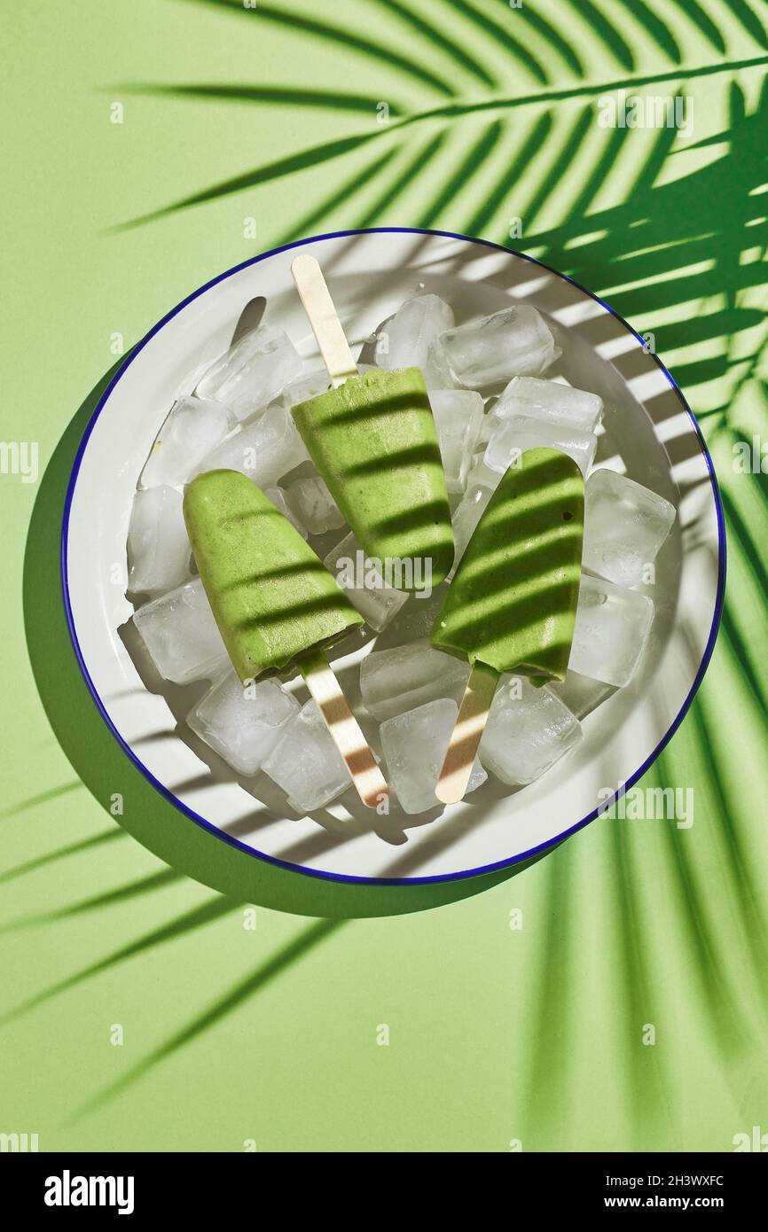 Gefrorener hausgemachter Pistazien-Popsicle in einer Schüssel mit Eis auf grünem Hintergrund. Erfrischender Fruchteis, gefrorener grüner Saft auf Stick. Top Vie Stockfoto