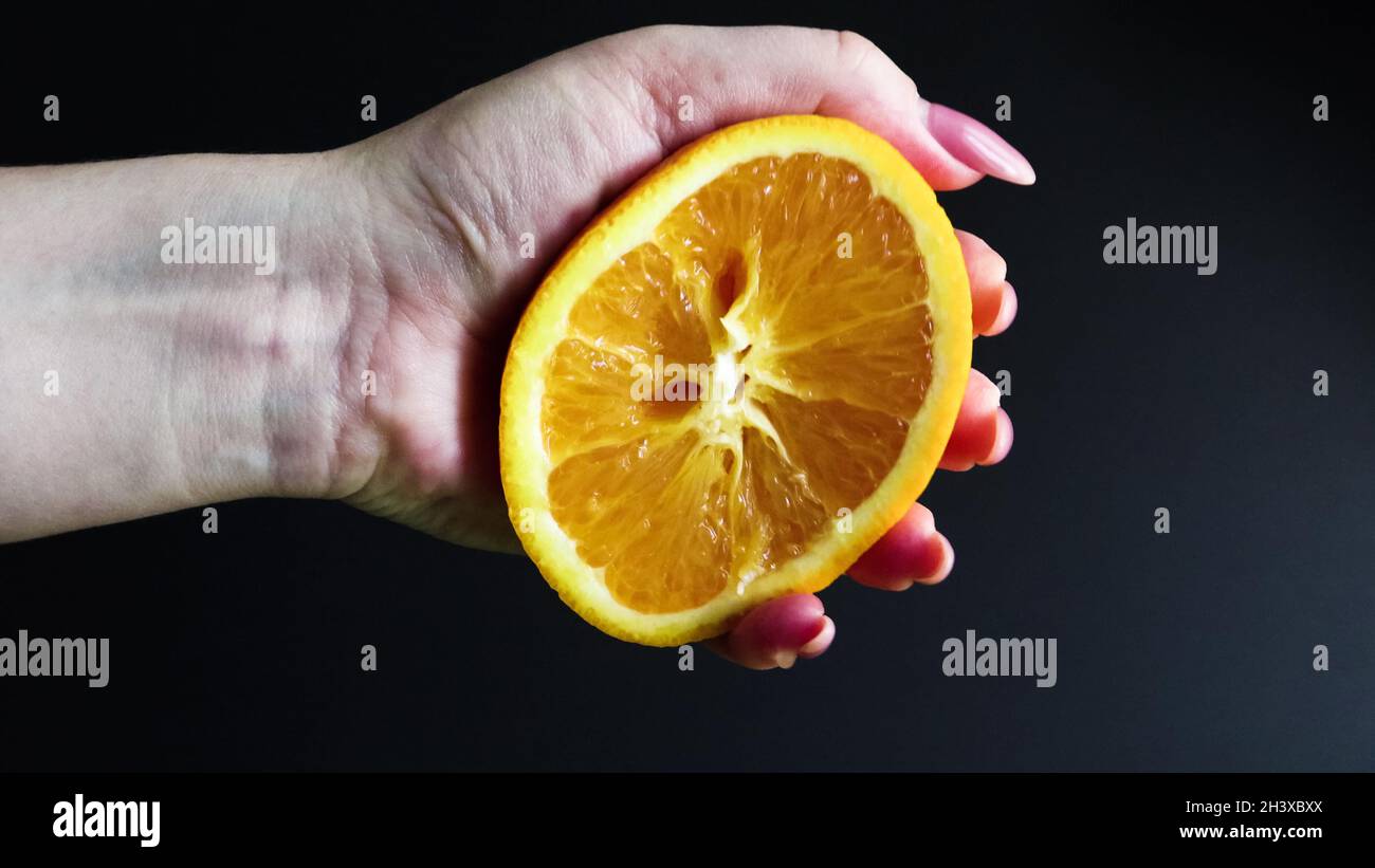 Eine weibliche Hand drückt die Hälfte einer frischen und saftigen Orange. Orangensaft aus Zitrusfrüchten tropft auf schwarzem Hintergrund aus Orangenfleisch. Th Stockfoto