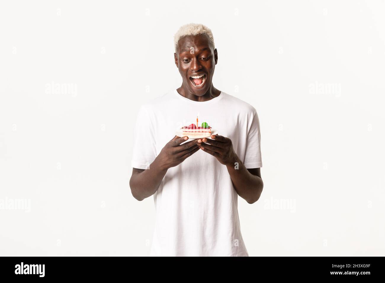Porträt eines hübschen, aufgeregten afroamerikanischen blonden Mannes, der amüsiert über den Geburtstagskuchen aussieht, lächelt und sich auf einer brennenden Kerze wünscht Stockfoto
