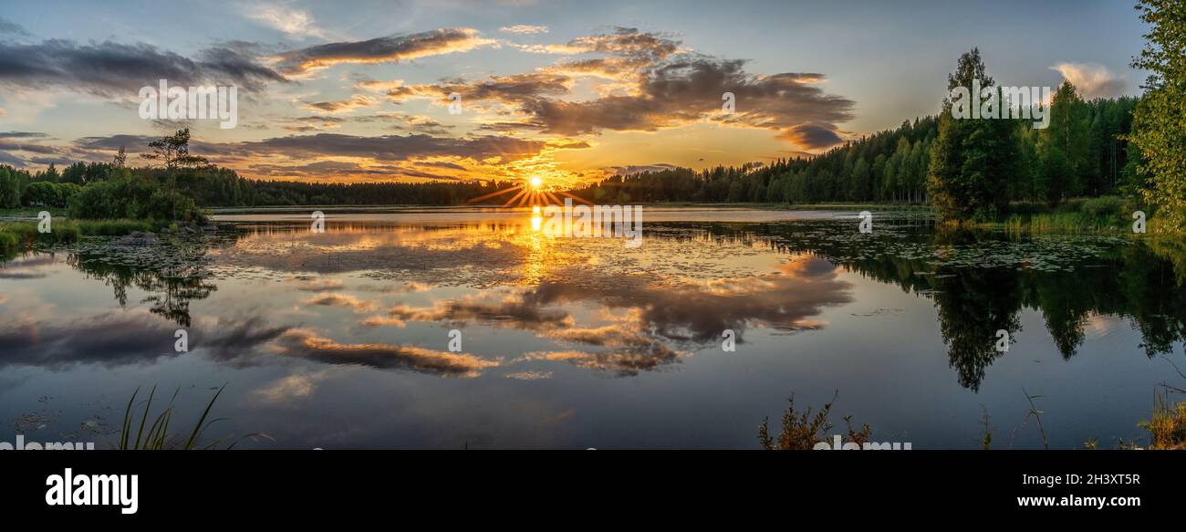 Ein Panorama eines farbenfrohen Sonnenuntergangs spiegelt sich in einer ruhigen Seenlandschaft mit grünem Wald und Schilf wider Stockfoto