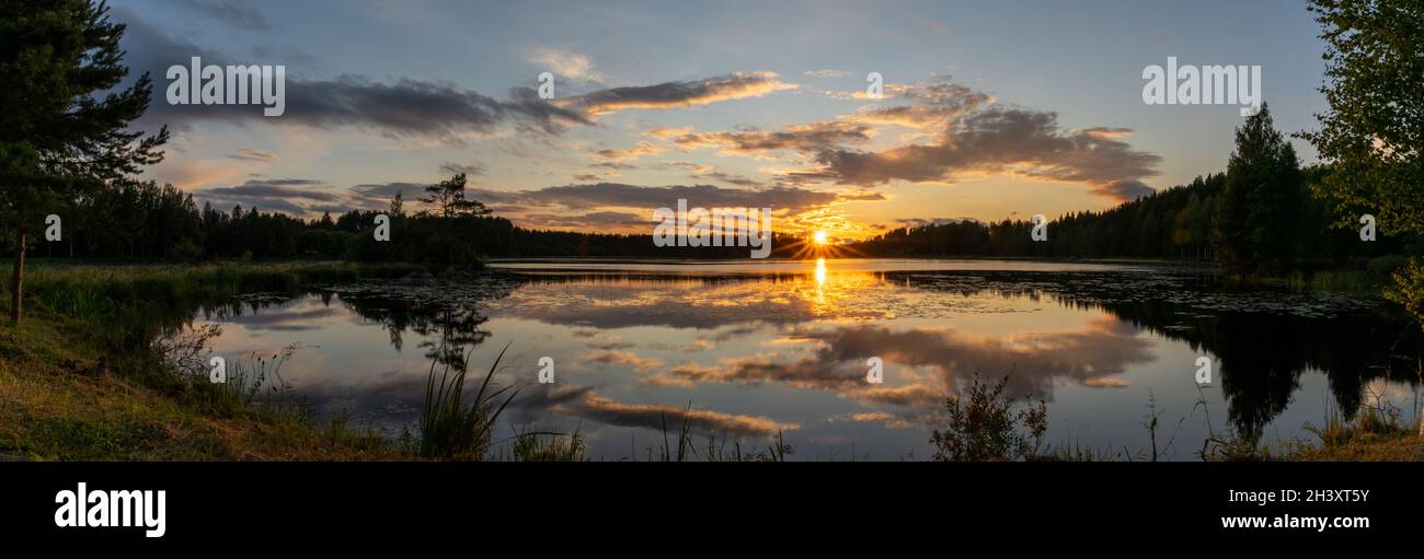 Ein Panorama eines farbenfrohen Sonnenuntergangs spiegelt sich in einer ruhigen Seenlandschaft mit grünem Wald und Schilf wider Stockfoto