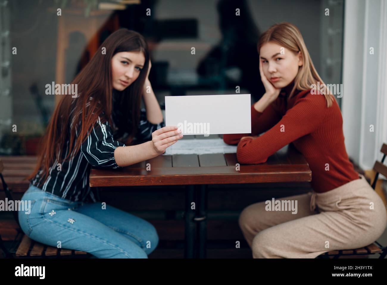 Frau hält weißes Papier in der Hand. Zwei Frauen unterhalten sich am Tisch im Straßencafé. Weißes leeres Vorlagenblatt mit leerem Platz. Stockfoto