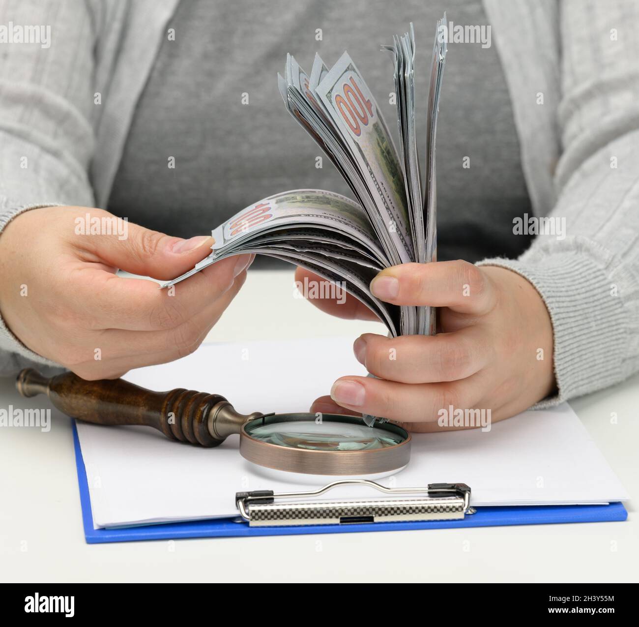 Ein Stapel von hundert Dollar Scheinen in der Hand einer Frau und eine hölzerne Lupe auf einem weißen Tisch. Budgetanalyse, Einkommen und Stockfoto