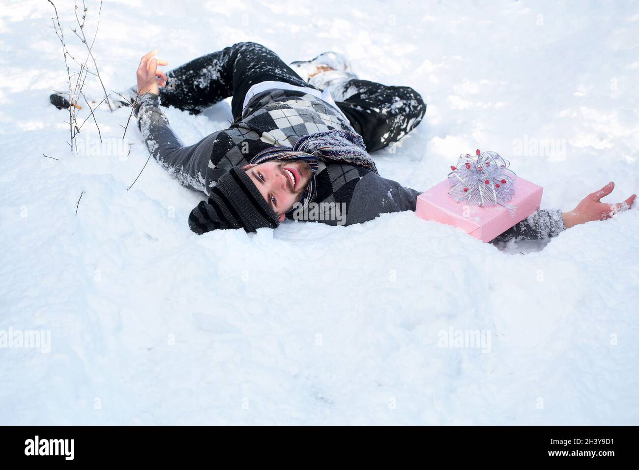 Crtazy Mann mit Geschenk-Box liegt auf Schnee. Weihnachtsgrüße und Weihnachtsgeschenke. Macho Homosexuell lächeln in Hut, Pullover und Schal am Wintertag Stockfoto