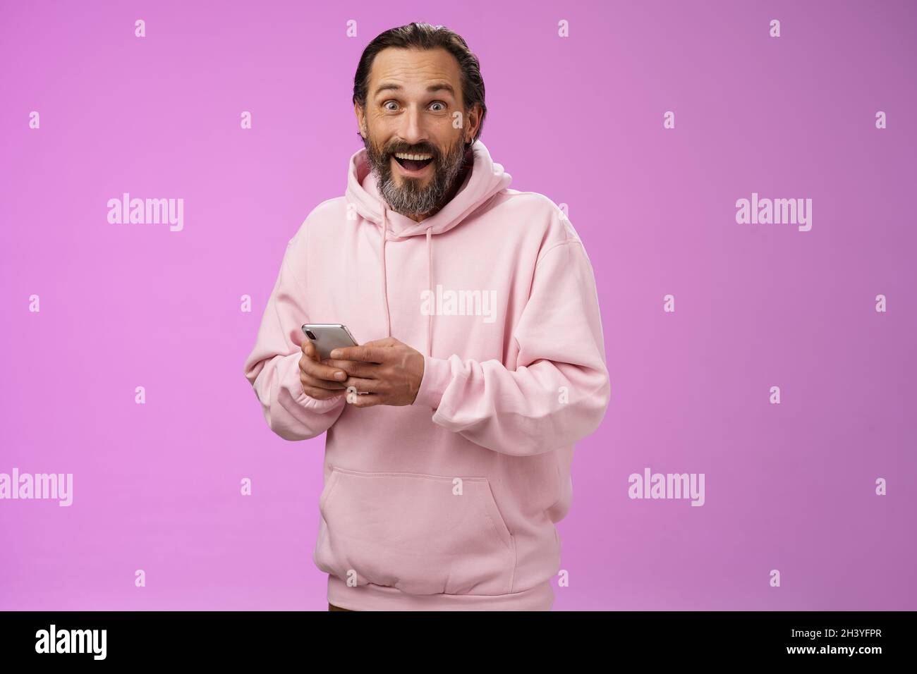 Amüsiert glücklich fasziniert erwachsenen reifen kaukasischen bärtigen Mann in rosa Hoodie keuchend beeindruckt halten Smartphone aussehen Kamera aston Stockfoto
