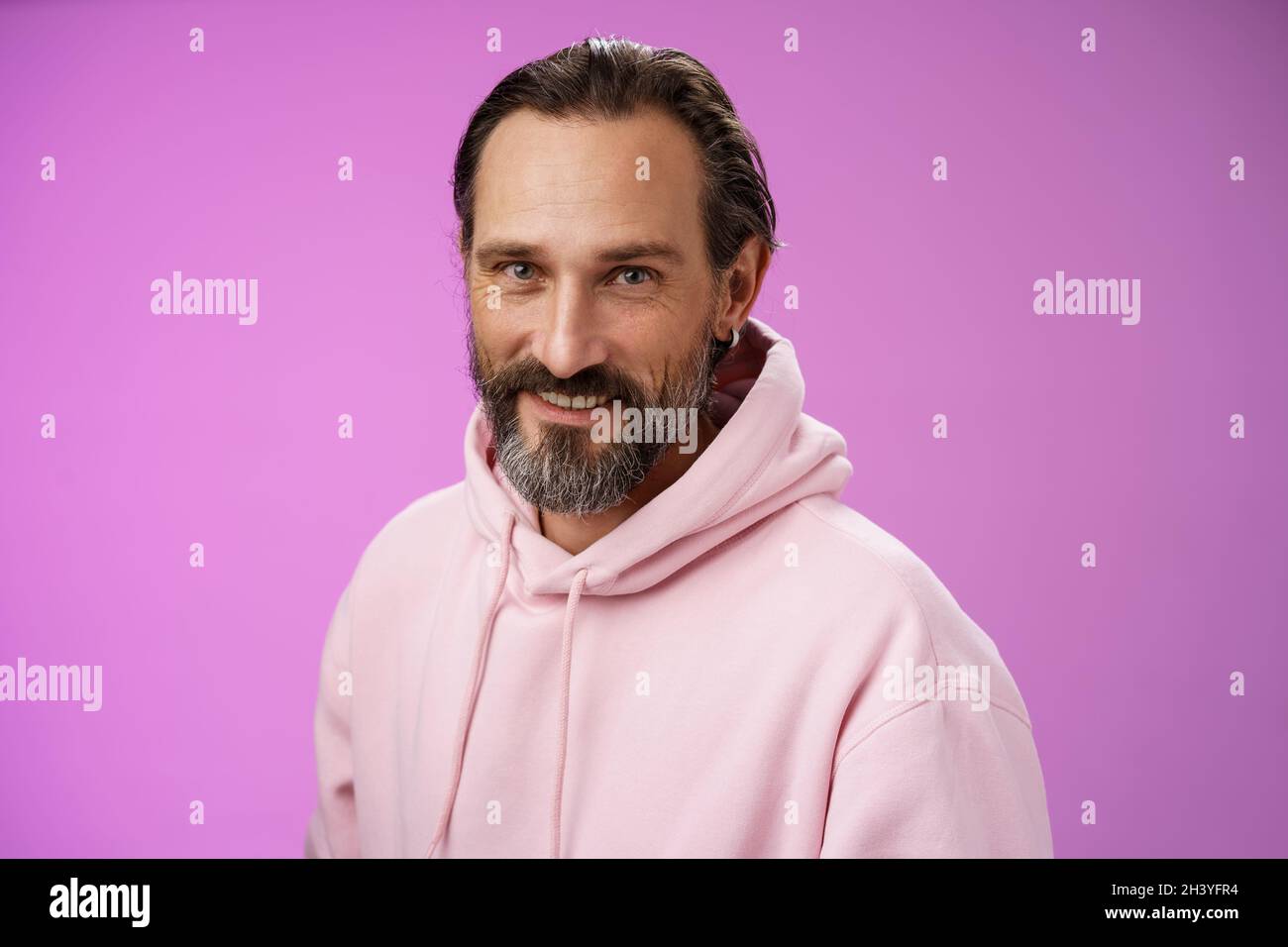 Charming verführerisch gut aussehend bärtig stilvolle Erwachsene männliche Modell Ohrring rosa Kapuzenpullover lächelnd erfreut Ausdruck Vertrauen Positivität Stockfoto