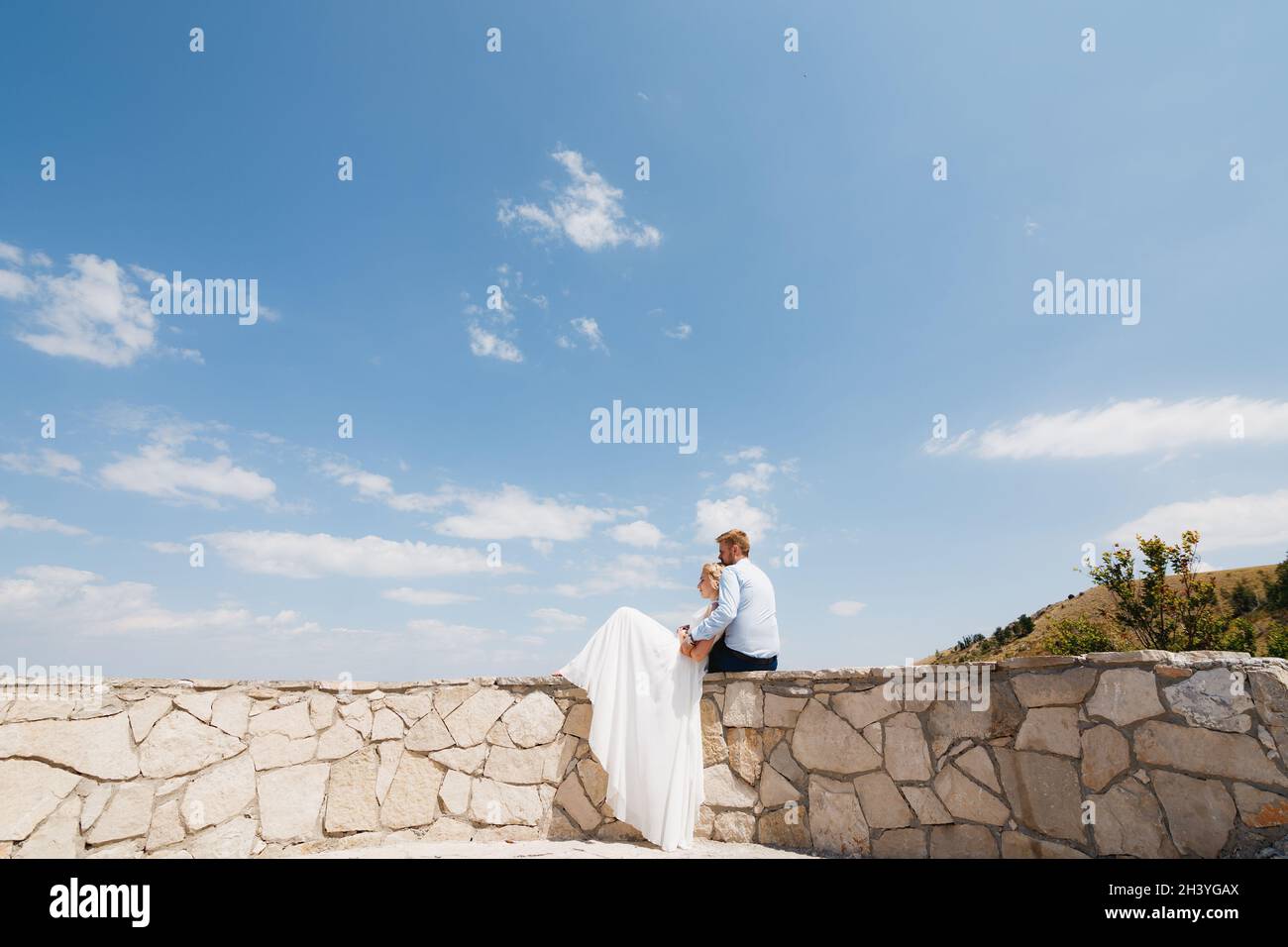 Braut und Bräutigam sitzen auf einem Steinzaun, die Braut lehnt sich an den Bräutigam, der Bräutigam umarmt sie sanft Stockfoto