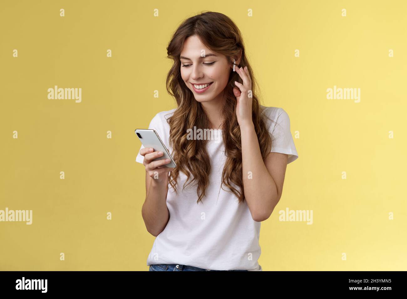 Zartes feminines, lockiges weibliches weißes T-Shirt mit Touch-Ohrstöpsel in Form des Ohrhörers mit lächelndem, fröhlich anmutendem Smartphone-Bildschirm Stockfoto