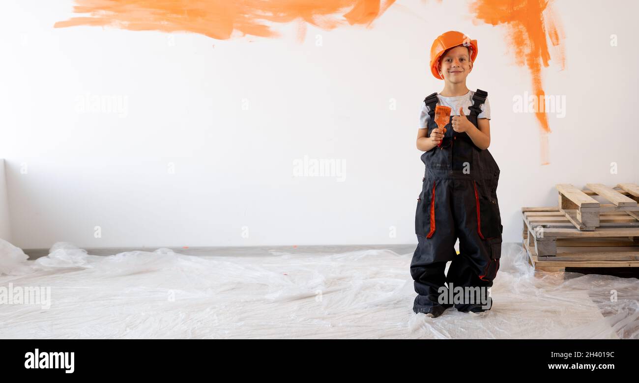 Schlüsselfertige Reparatur. Kleiner Junge in einem Helm und Overalls mit einem Pinsel auf dem Hintergrund einer weißen Wand mit oranger Farbe. Neues Haus renoviert Stockfoto