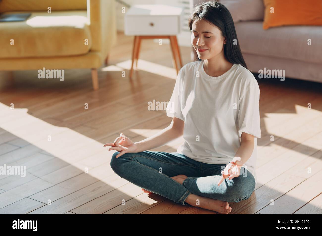 Asiatische Frau, die Yoga und Zen wie Meditation Lotuspose in lässiger Kleidung im Wohnzimmer Wohnung mit natürlichem Sonnenlicht Stockfoto