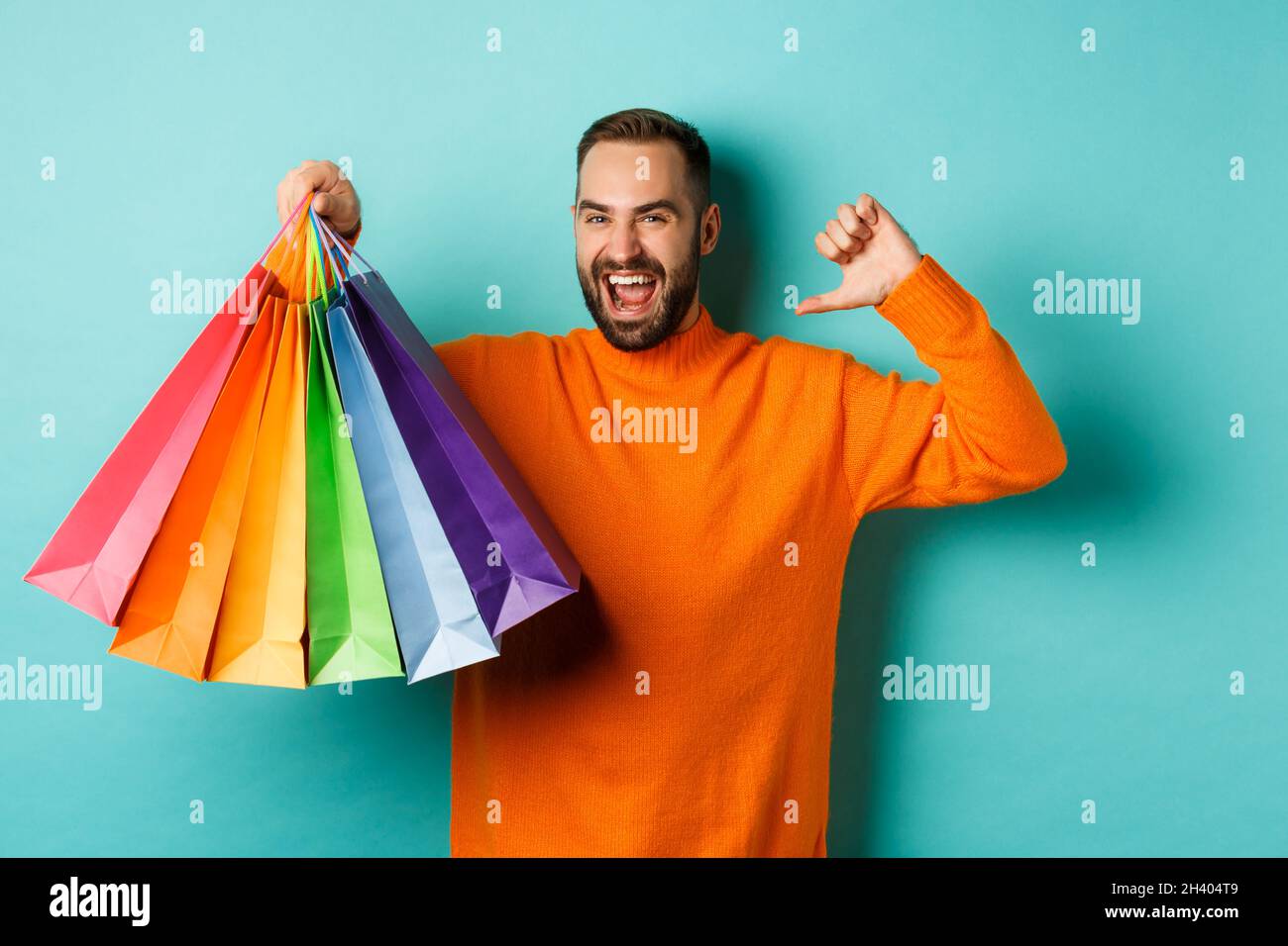 Hübscher bärtiger Mann in orangefarbenem Pullover, der Geschenke kauft, auf sich zeigt und Einkaufstaschen zeigt und über blauem Backgrou steht Stockfoto