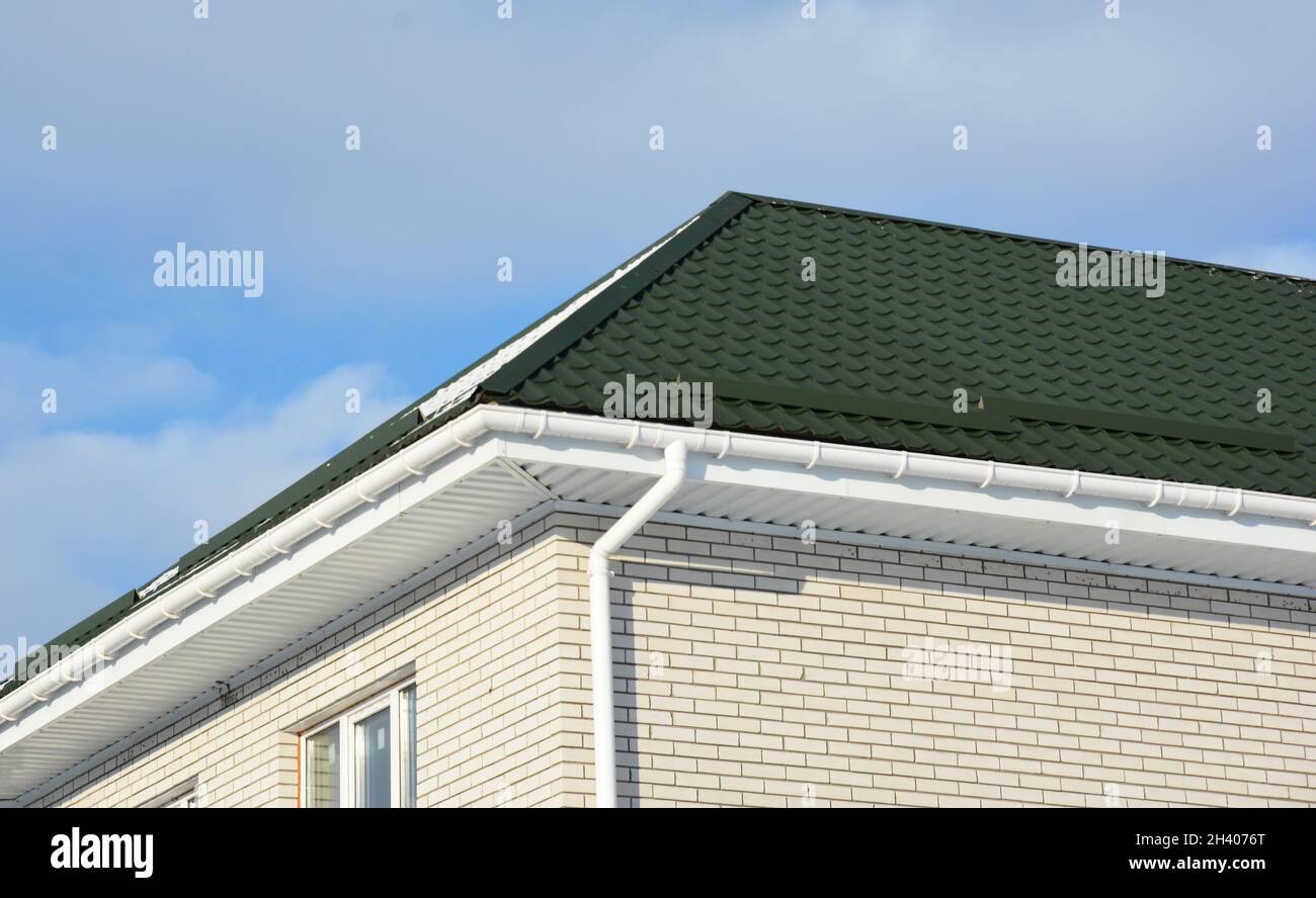 Ein Backsteinhaus mit einem grünen Metalldach, weißem Kunststoffsoffit und Faszien und einer Dachrinnenleitung, Downauslauf. Stockfoto