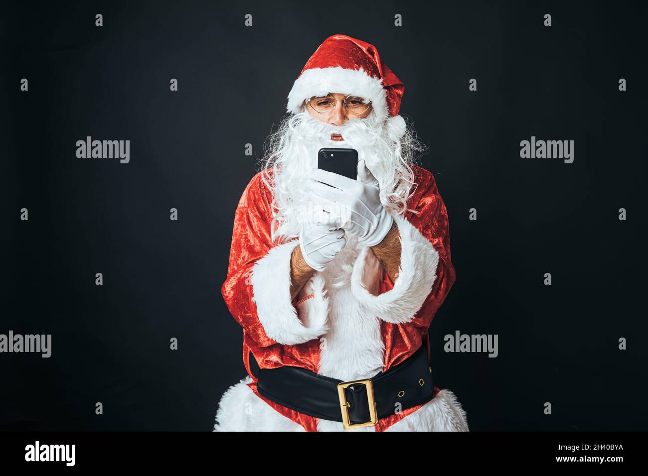 Mann, der als Weihnachtsmann gekleidet war und auf sein Mobiltelefon schaute, auf schwarzem Hintergrund. Konzept von Weihnachten, Weihnachtsmann, Geschenke, Feier. Stockfoto