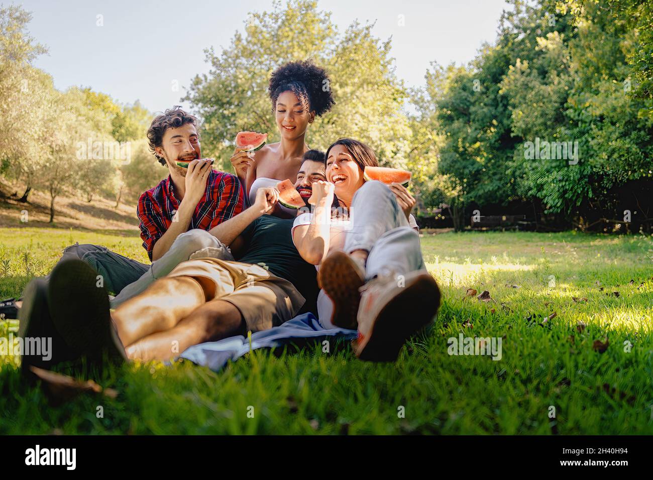 Eine Gruppe von glücklichen jungen Menschen, die beim Picknick auf dem Gras sitzen und Spaß daran haben, Wassermelone zu essen. Gefiltertes Bild mit lebendigen warmen Farben. Stockfoto