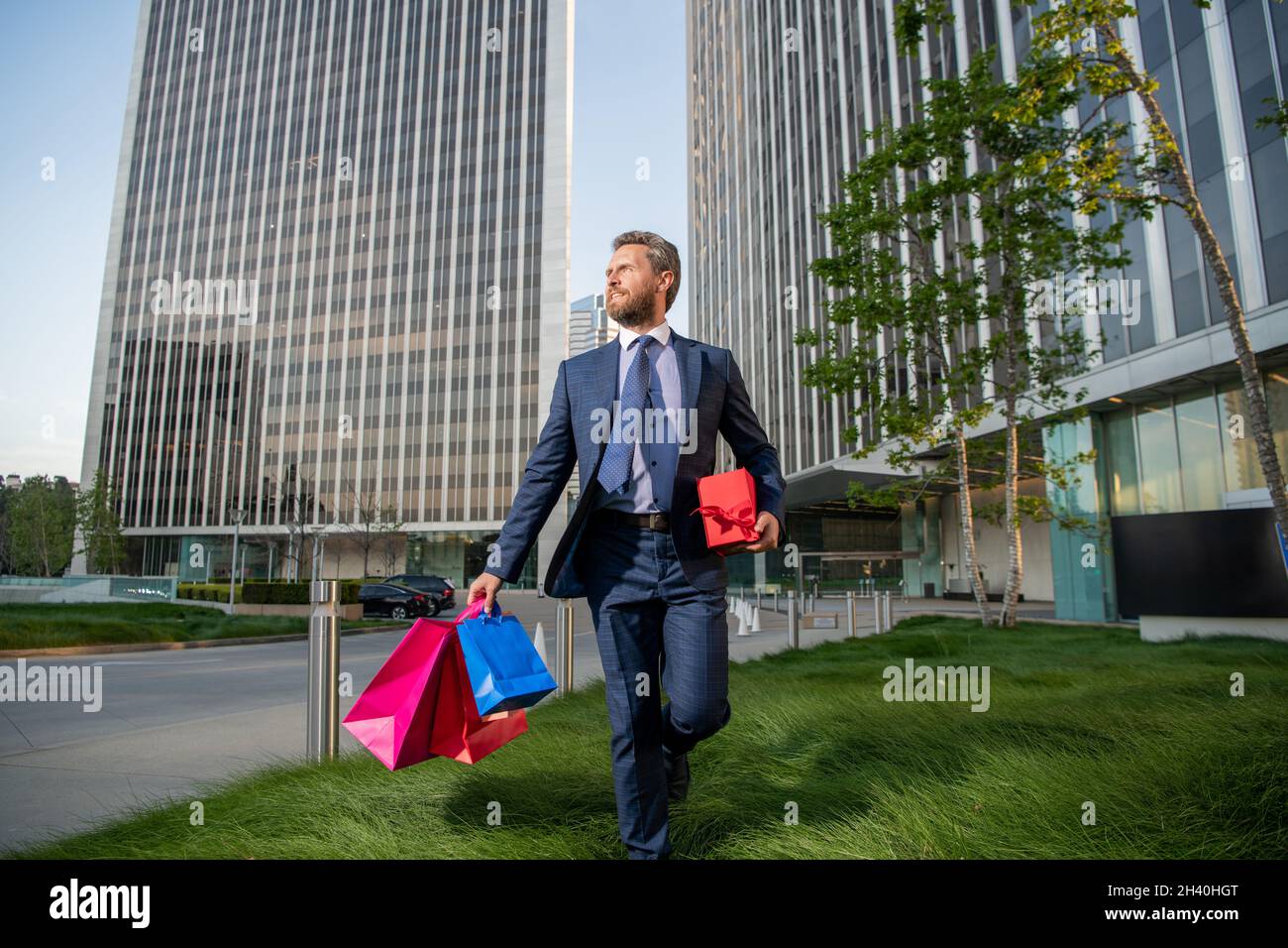 Glücklicher Unternehmer in Anzug hält Einkaufstasche und geschenkbox vor dem Büro, Herrentag. Stockfoto
