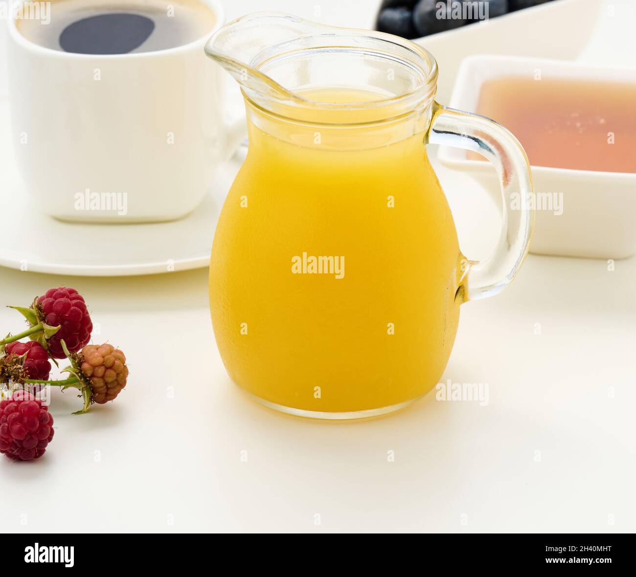 Frisch gepresster Saft in einer transparenten Glasdekanter, eine Tasse Kaffee, Honig auf einem weißen Tisch. Frühstück am Morgen Stockfoto