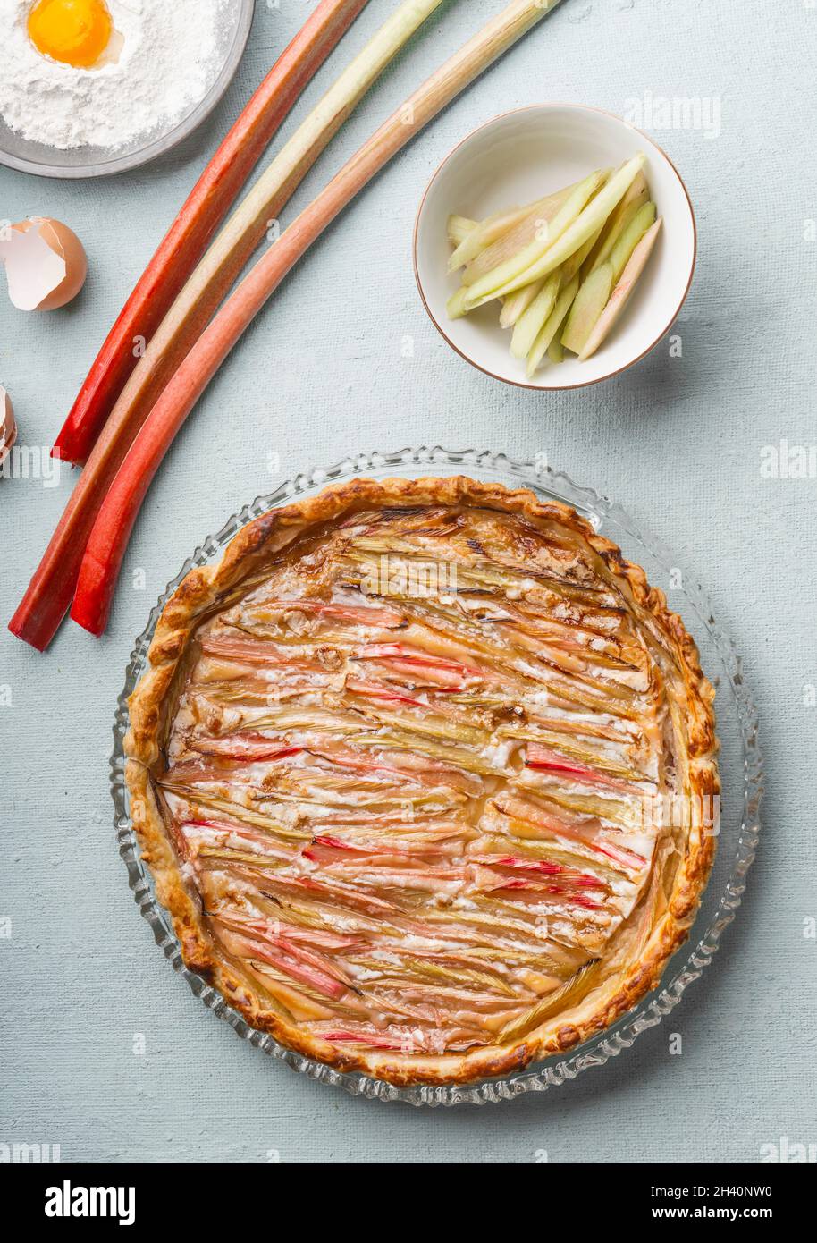 Hausgemachter Rhabarberkuchen auf grauem Küchentisch mit Rhabarber, Mehl mit Eigelb, Eierschale und Schüssel. Saisonales Backen zu Hause. Draufsicht. Stockfoto