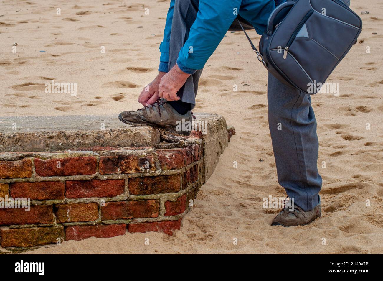 Ein älterer kaukasischer Mann mit einer Tasche ruht seinen Fuß auf einer Ziegelwand, die an einem Sandstrand versunken ist, um seine Schnürsenkel zu binden Stockfoto