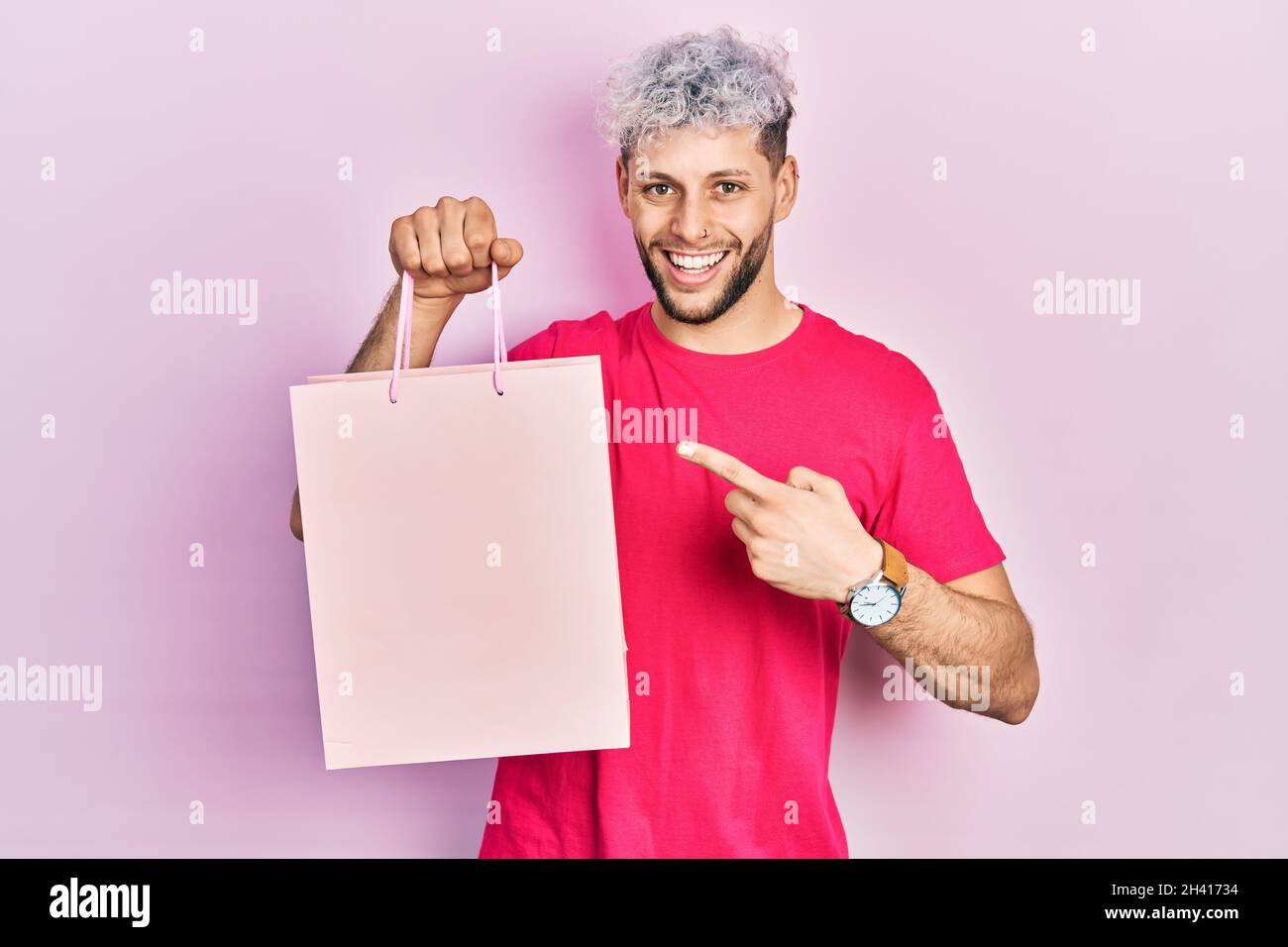 Junger hispanischer Mann mit modernen gefärbten Haaren hält Einkaufstasche lächelnd glücklich zeigt mit Hand und Finger Stockfoto