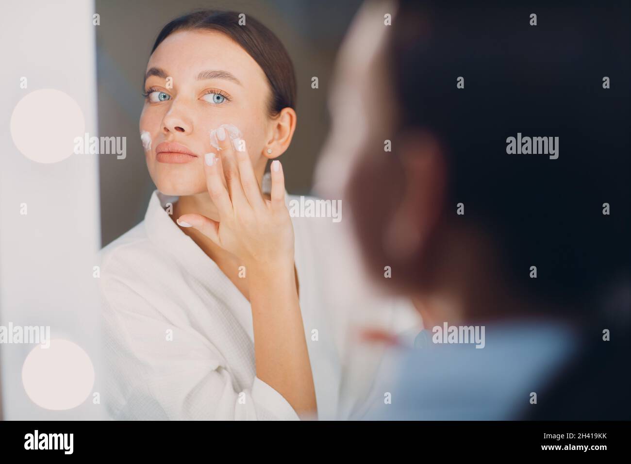 Junge Frau, die am Spiegel kosmetische weiße Creme auf ihr Gesicht appied Stockfoto