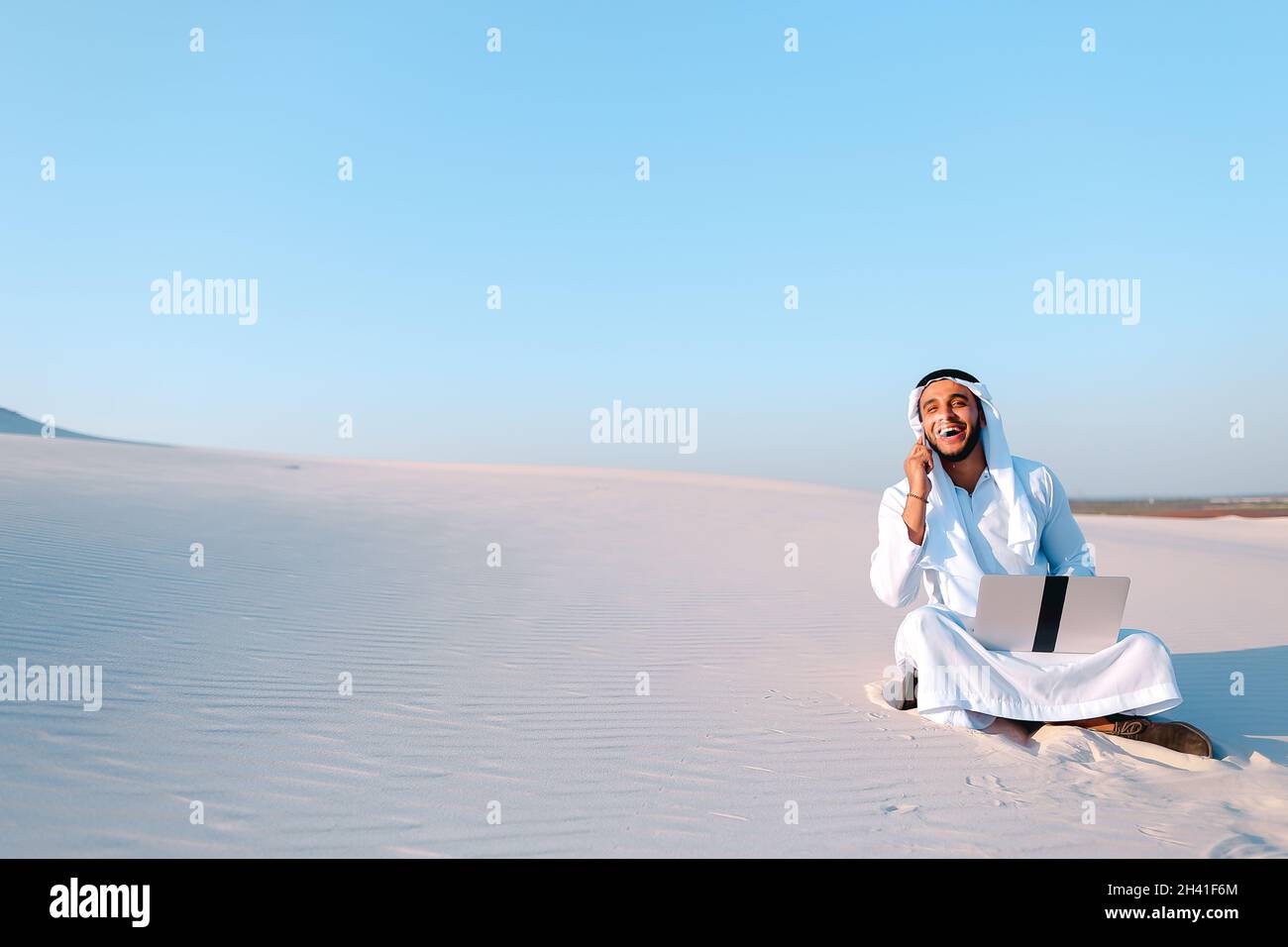 Der erfolgreiche muslimische Architekt kommuniziert am heißen Morgen telefonisch mit dem Kunden, der auf Sand mit einem Laptop in der großen Wüste sitzt. Stockfoto