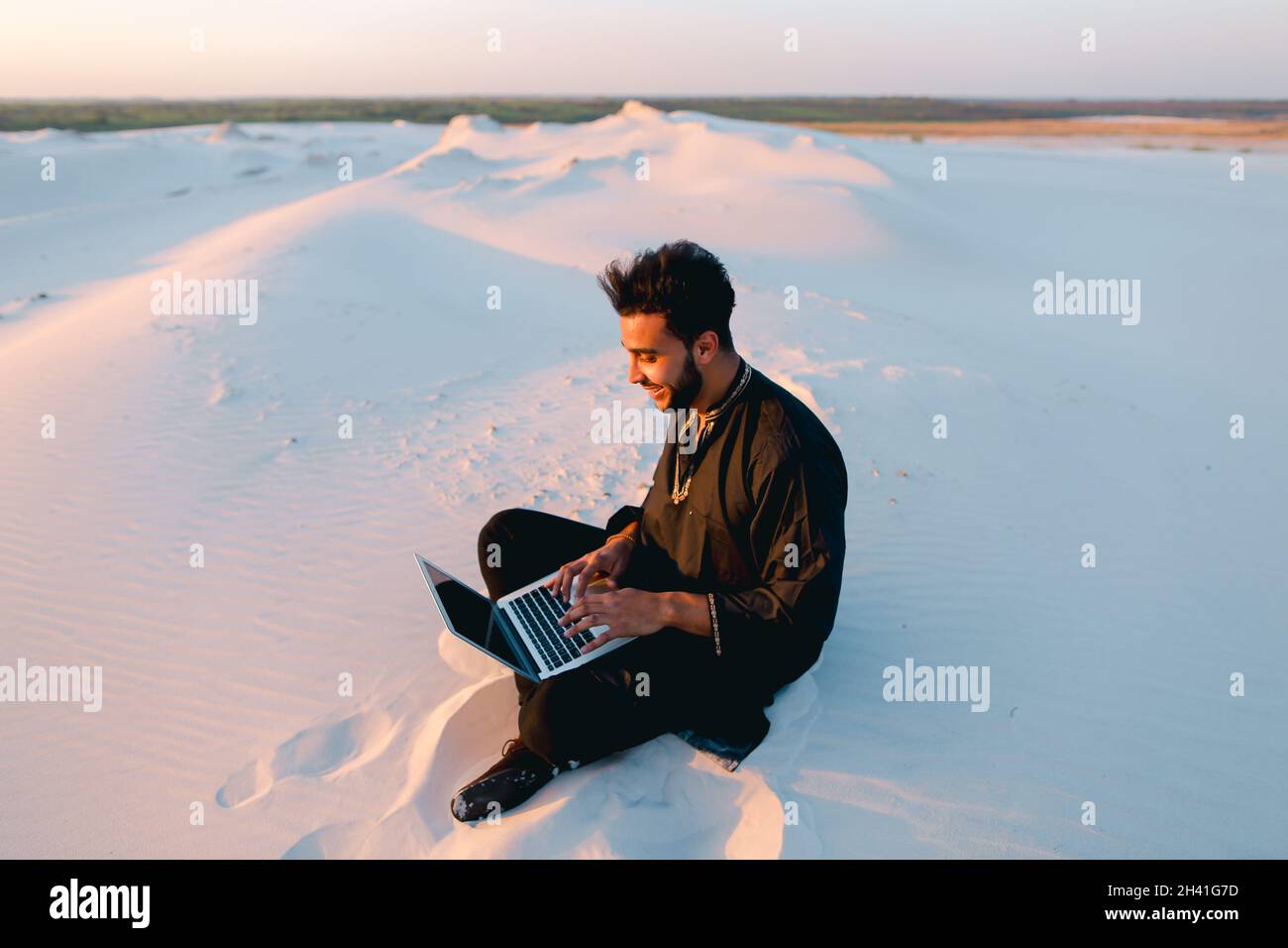 Ein hübscher junger arabischer Mann schreibt mit einem Laptop über das Internet und sitzt bei Sonnenuntergang auf Sand inmitten einer sandigen Wüste. Stockfoto