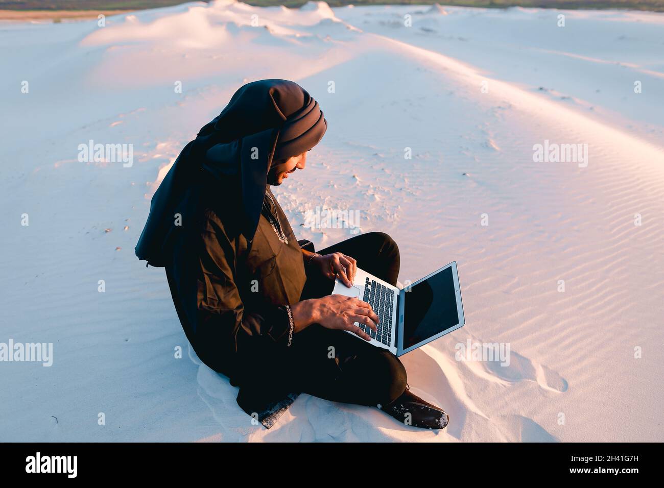 Der gebildete arabische Student verwendet einen Laptop und arbeitet am warmen Sommerabend auf Sand inmitten von Sanddünen der Wüste. Stockfoto