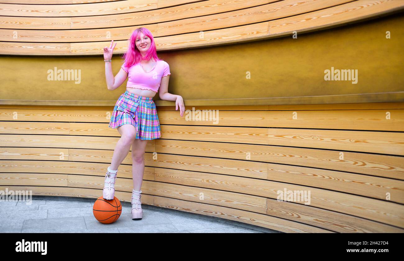 Ein schönes Mädchen mit rosa Haaren posiert vor der Wand - Weibliche Millennial steht im Urbanity Werbebereich mit dem Fuß auf der Sonnenbass ruhen Stockfoto