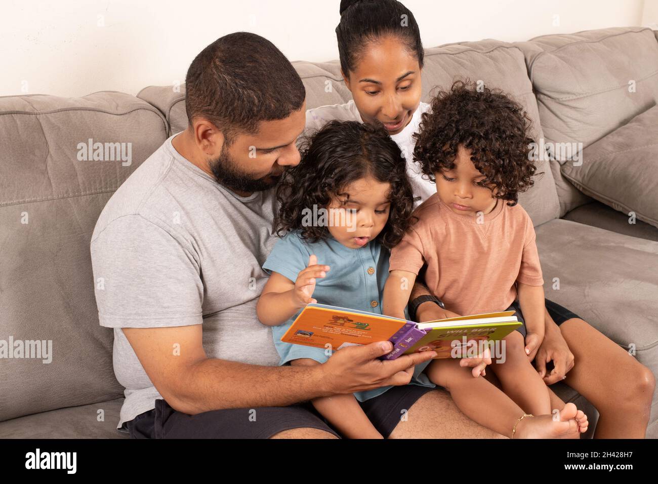2 Jahre alte brüderliche Zwillinge, Junge und Mädchen, die mit den Eltern auf der Couch sitzen, lesen aus den Bordbüchern Stockfoto